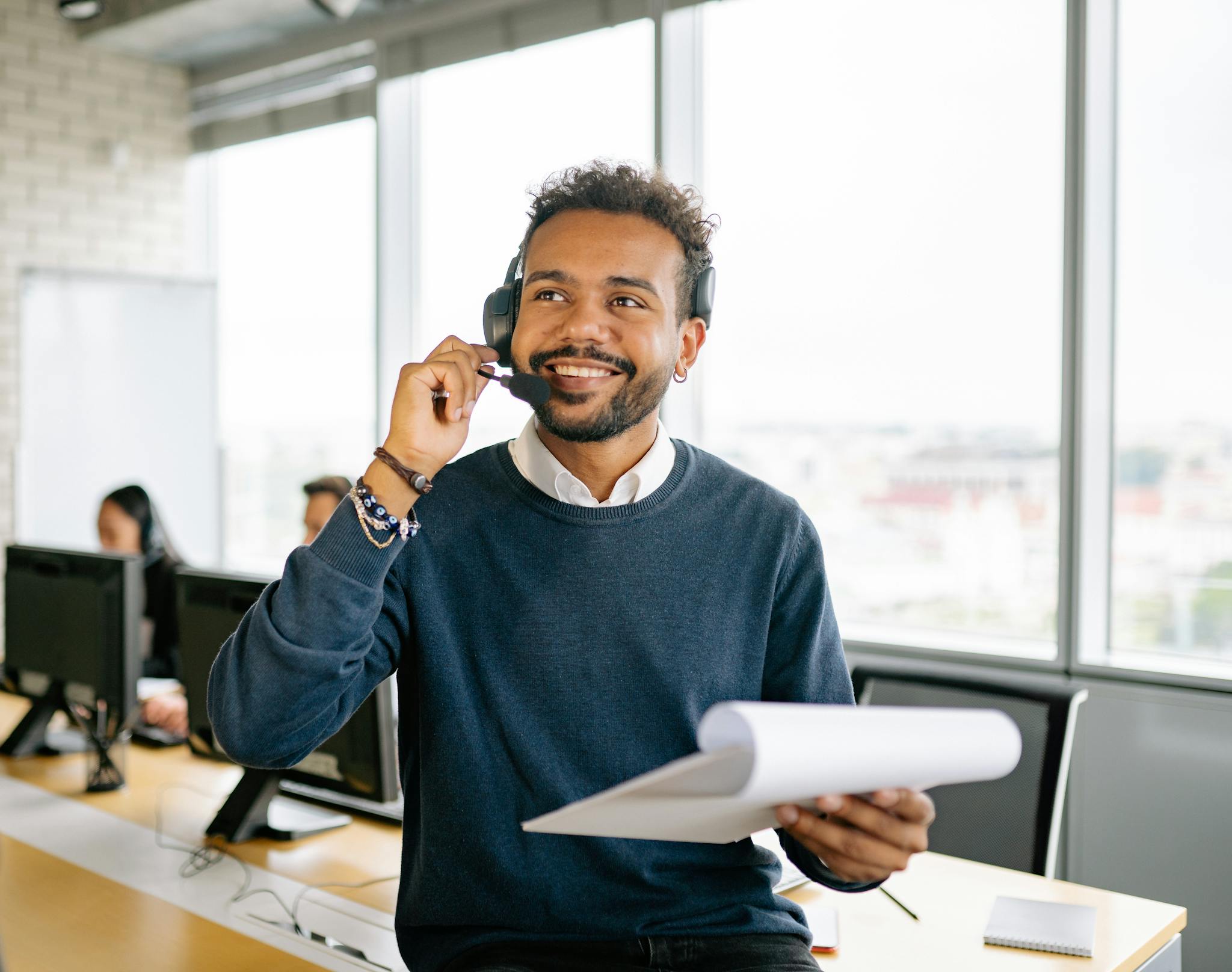 A cheerful man wearing a headset and holding a clipboard at a modern office workspace.